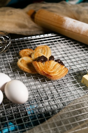 A baking scene features flaky pastries with chocolate, resting on a cooling rack dusted with flour. Nearby, fresh eggs, a block of butter, a whisk, and a wooden rolling pin are visible, suggesting a preparation setting. The overall atmosphere conveys a sense of home baking, warmth, and deliciousness.