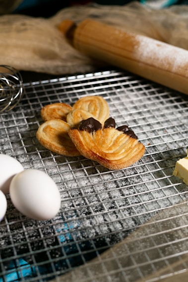 A baking scene features flaky pastries with chocolate, resting on a cooling rack dusted with flour. Nearby, fresh eggs, a block of butter, a whisk, and a wooden rolling pin are visible, suggesting a preparation setting. The overall atmosphere conveys a sense of home baking, warmth, and deliciousness.