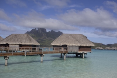 Overwater bungalows with thatched roofs stand on stilts above a clear turquoise sea. A wooden walkway connects the bungalows, and in the background, there are lush green mountains under a sky dotted with clouds.