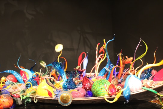A vibrant and intricate glass sculpture featuring an array of twisting, colorful glass elements. The sculpture includes spirals, orbs, and elongated shapes in a variety of bright colors including blue, red, yellow, and green. These elements are artistically arranged in a large bowl-like structure set against a dark background, highlighting the vividness of the glass.
