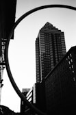 A high-contrast black and white photograph of an urban scene featuring a tall, modern skyscraper with a triangular top in the center. The image captures a unique perspective with a large circular structure partially surrounding the buildings. The foreground and background buildings have repetitive window patterns and clean architectural lines.
