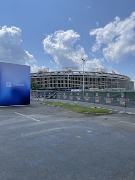 A large stadium with a distinctive oval design is visible behind a fence with banners labeled 'Electrify Expo.' The foreground features an asphalt parking lot with a blue billboard displaying the Toyota logo on the left. Large, fluffy clouds punctuate the bright blue sky above.