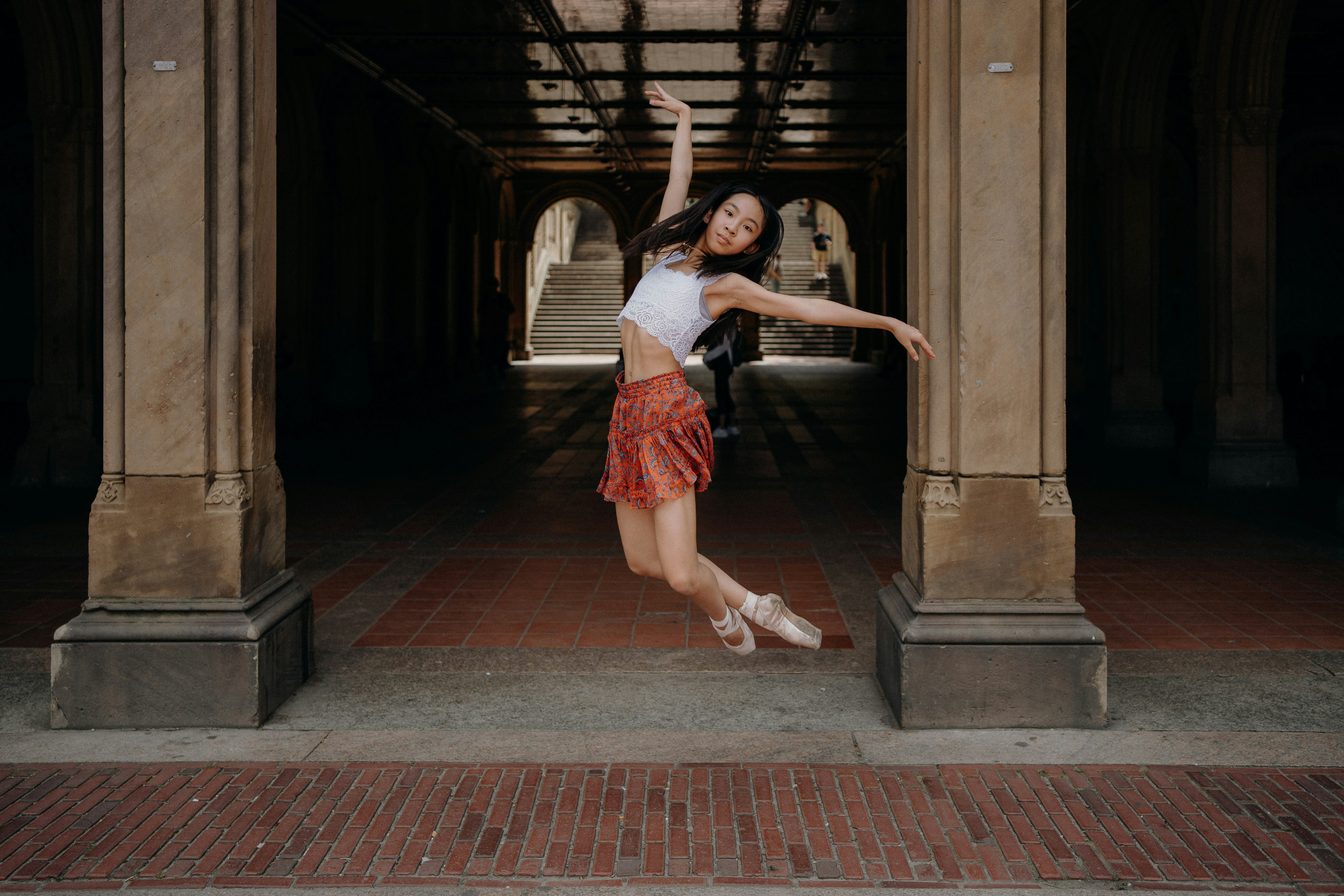 A dancer gracefully leaps in the air under a large archway. She wears a white top and a red skirt with ballet shoes, striking a pose with arms outstretched. The background features stone pillars and a tiled floor, with light filtering through, creating a dramatic effect.
