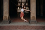 A dancer gracefully leaps in the air under a large archway. She wears a white top and a red skirt with ballet shoes, striking a pose with arms outstretched. The background features stone pillars and a tiled floor, with light filtering through, creating a dramatic effect.