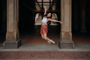 A dancer gracefully leaps in the air under a large archway. She wears a white top and a red skirt with ballet shoes, striking a pose with arms outstretched. The background features stone pillars and a tiled floor, with light filtering through, creating a dramatic effect.