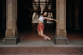 A dancer gracefully leaps in the air under a large archway. She wears a white top and a red skirt with ballet shoes, striking a pose with arms outstretched. The background features stone pillars and a tiled floor, with light filtering through, creating a dramatic effect.