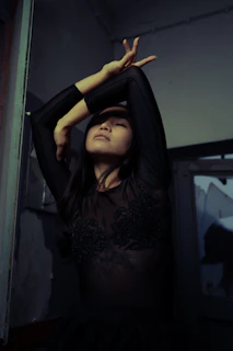 A woman with long dark hair wears a black, intricately designed, semi-transparent top. Her eyes are closed, and her arms are gracefully positioned above her head, creating an elegant pose against a dimly lit, indoor background.