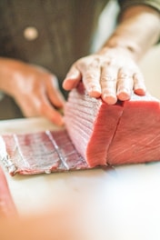 A hand is holding a large piece of raw tuna on a cutting board, with another hand using a knife to slice it into thinner pieces. The focus is on the freshness and texture of the fish.