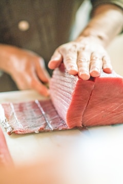A hand is holding a large piece of raw tuna on a cutting board, with another hand using a knife to slice it into thinner pieces. The focus is on the freshness and texture of the fish.