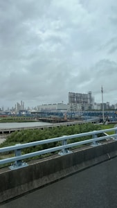 An industrial complex with large structures and equipment, possibly a factory or processing plant, set against a cloudy sky. A bridge with railings is in the foreground, and there is lush greenery below.