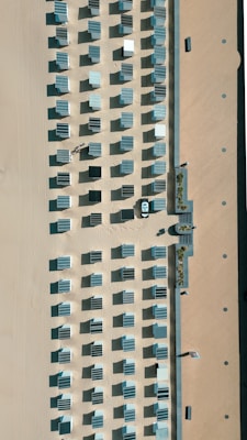 Aerial view of a beach area with neatly arranged rows of cabanas casting long shadows on the sand. The cabanas are uniformly spaced, creating a grid pattern accentuated by the sunlight. Visible pathways and a small building with steps lead up to the beach. A solitary figure is visible among the cabanas.