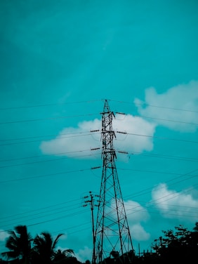 A tall metal electricity pylon stands against a bright teal sky with scattered clouds. Power lines extend from it in various directions. Below, there are silhouettes of palm trees and foliage.