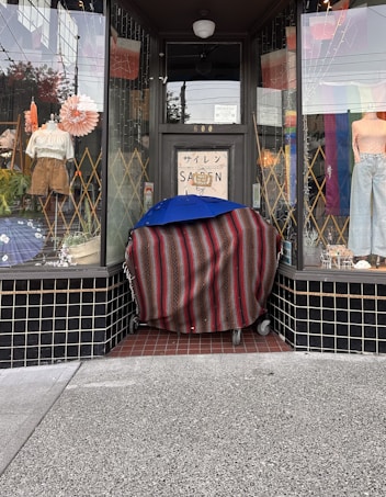 A storefront displays various items, with mannequins showcasing clothing in the windows. A cloth-covered object on wheels is placed in front, topped with a blue umbrella. The entrance door has signage in both English and Japanese scripts.
