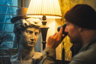 A classical marble bust wears a stylish hat, positioned beside a vintage-style lamp casting a warm glow. A person in a beanie focuses intently through a monocular device.