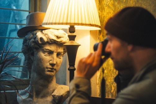 A classical marble bust wears a stylish hat, positioned beside a vintage-style lamp casting a warm glow. A person in a beanie focuses intently through a monocular device.