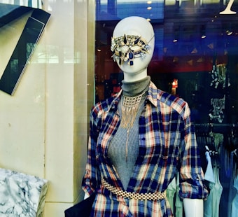 A mannequin is dressed in a stylish outfit featuring a plaid shirt layered over a gray turtleneck. It wears an elaborate, decorative face mask comprised of blue and gold elements. A gold chain belt cinches the waist, and a statement necklace adds to the ensemble. The background includes a store setting with clothing racks.