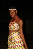A person is wearing traditional colorful attire with geometric patterns. The outfit consists of a top and a matching skirt, adorned with vibrant yellow, pink, and blue design. The individual has facial makeup and a white beaded headband, standing against a dark background.
