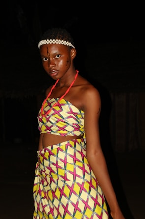 A person is wearing traditional colorful attire with geometric patterns. The outfit consists of a top and a matching skirt, adorned with vibrant yellow, pink, and blue design. The individual has facial makeup and a white beaded headband, standing against a dark background.