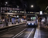 A tram is stationed at a well-lit urban platform during nighttime. The tram's headlights are on, and there is a visible public transportation logo. Passengers are visible inside the tram, and a person is walking along the near-empty platform. Overhead, a metro train is crossing a bridge, illuminating the surrounding area with artificial lights. The setting is an urban environment with infrastructure such as railings, platforms, and digital displays.