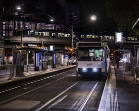 A tram is stationed at a well-lit urban platform during nighttime. The tram's headlights are on, and there is a visible public transportation logo. Passengers are visible inside the tram, and a person is walking along the near-empty platform. Overhead, a metro train is crossing a bridge, illuminating the surrounding area with artificial lights. The setting is an urban environment with infrastructure such as railings, platforms, and digital displays.