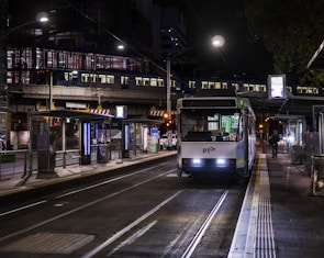 A tram is stationed at a well-lit urban platform during nighttime. The tram's headlights are on, and there is a visible public transportation logo. Passengers are visible inside the tram, and a person is walking along the near-empty platform. Overhead, a metro train is crossing a bridge, illuminating the surrounding area with artificial lights. The setting is an urban environment with infrastructure such as railings, platforms, and digital displays.