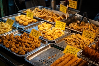 An assortment of fried foods displayed in metal trays, including chicken nuggets, chicken gizzard, chicken popcorn, and crispy fish skin. Each tray is labeled with a yellow sign, and the selection includes both chicken and fish items. The food is golden brown and appears to be well seasoned.