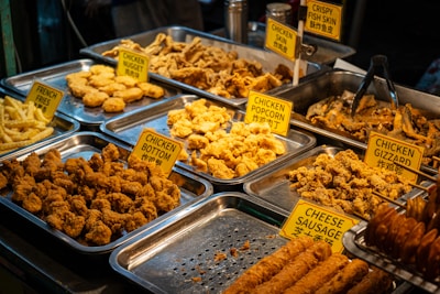 An assortment of fried foods displayed in metal trays, including chicken nuggets, chicken gizzard, chicken popcorn, and crispy fish skin. Each tray is labeled with a yellow sign, and the selection includes both chicken and fish items. The food is golden brown and appears to be well seasoned.
