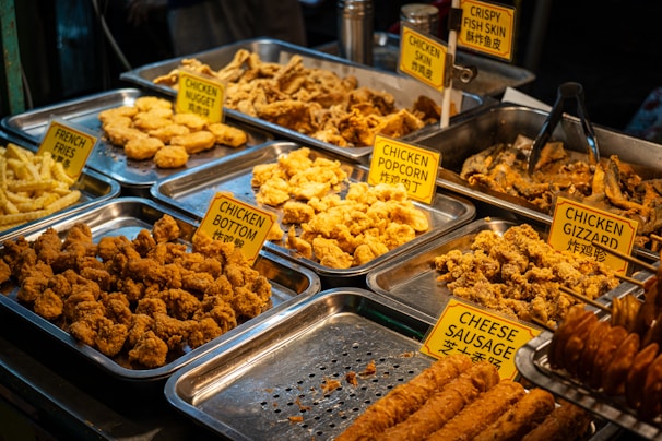 An assortment of fried foods displayed in metal trays, including chicken nuggets, chicken gizzard, chicken popcorn, and crispy fish skin. Each tray is labeled with a yellow sign, and the selection includes both chicken and fish items. The food is golden brown and appears to be well seasoned.