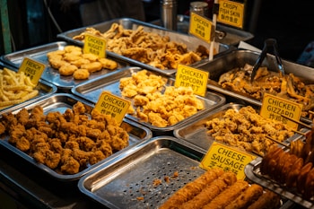 An assortment of fried foods displayed in metal trays, including chicken nuggets, chicken gizzard, chicken popcorn, and crispy fish skin. Each tray is labeled with a yellow sign, and the selection includes both chicken and fish items. The food is golden brown and appears to be well seasoned.