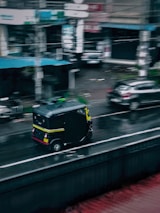 A black and yellow auto rickshaw is driving on a wet road in an urban setting. The surroundings include parked cars and buildings with blurry signage, suggesting a busy city environment on a rainy day.