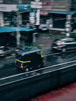 A black and yellow auto rickshaw is driving on a wet road in an urban setting. The surroundings include parked cars and buildings with blurry signage, suggesting a busy city environment on a rainy day.