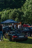 A group of people gather around various cars parked on a grassy area with a forested backdrop. A black convertible with a visible license plate is prominently displayed in the foreground. The event appears to be a casual car meet, with a canopy tent visible in the background providing shade.