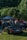 A group of people gather around various cars parked on a grassy area with a forested backdrop. A black convertible with a visible license plate is prominently displayed in the foreground. The event appears to be a casual car meet, with a canopy tent visible in the background providing shade.