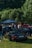 A group of people gather around various cars parked on a grassy area with a forested backdrop. A black convertible with a visible license plate is prominently displayed in the foreground. The event appears to be a casual car meet, with a canopy tent visible in the background providing shade.