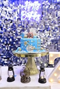 A blue birthday cake with gold and white decorations sits on a gold cake stand. The cake features two bear figurines on top, along with stars, clouds, and a ladder. The cake has the date 29th July 2023 written on it. In the background, there is a shimmering, blue sequined curtain with a neon sign that reads 'Happy Birthday'. Surrounding the cake are small decorative lanterns and a birdcage.