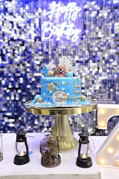 A blue birthday cake with gold and white decorations sits on a gold cake stand. The cake features two bear figurines on top, along with stars, clouds, and a ladder. The cake has the date 29th July 2023 written on it. In the background, there is a shimmering, blue sequined curtain with a neon sign that reads 'Happy Birthday'. Surrounding the cake are small decorative lanterns and a birdcage.