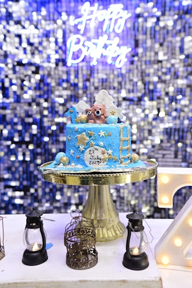 A blue birthday cake with gold and white decorations sits on a gold cake stand. The cake features two bear figurines on top, along with stars, clouds, and a ladder. The cake has the date 29th July 2023 written on it. In the background, there is a shimmering, blue sequined curtain with a neon sign that reads 'Happy Birthday'. Surrounding the cake are small decorative lanterns and a birdcage.
