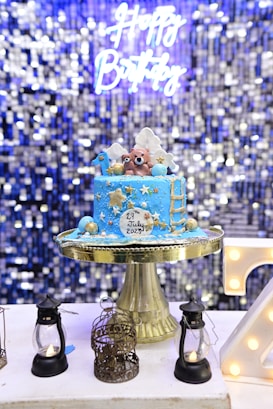 A blue birthday cake with gold and white decorations sits on a gold cake stand. The cake features two bear figurines on top, along with stars, clouds, and a ladder. The cake has the date 29th July 2023 written on it. In the background, there is a shimmering, blue sequined curtain with a neon sign that reads 'Happy Birthday'. Surrounding the cake are small decorative lanterns and a birdcage.