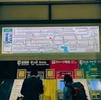 A train station ticket vending area displays a comprehensive rail network map above several automated ticket machines. People use the machines, and the map includes various colorful lines and text indicating the routes.