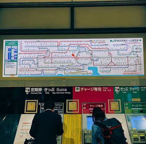 A train station ticket vending area displays a comprehensive rail network map above several automated ticket machines. People use the machines, and the map includes various colorful lines and text indicating the routes.