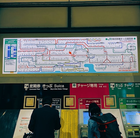 A train station ticket vending area displays a comprehensive rail network map above several automated ticket machines. People use the machines, and the map includes various colorful lines and text indicating the routes.