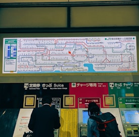 A train station ticket vending area displays a comprehensive rail network map above several automated ticket machines. People use the machines, and the map includes various colorful lines and text indicating the routes.