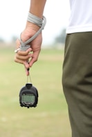 A person is holding a stopwatch in their left hand with a strap wrapped around their wrist. The background is an out-of-focus field or grass area. The person is wearing a white shirt and dark green sports pants.