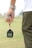 A person is holding a stopwatch in their left hand with a strap wrapped around their wrist. The background is an out-of-focus field or grass area. The person is wearing a white shirt and dark green sports pants.