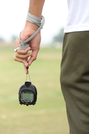 A person is holding a stopwatch in their left hand with a strap wrapped around their wrist. The background is an out-of-focus field or grass area. The person is wearing a white shirt and dark green sports pants.