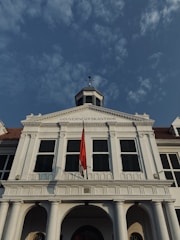 A historic white building with a tall structure, featuring an archway and large windows with green shutters. The building's facade is adorned with columns and a red and white flag is prominently displayed at the front. The sky above is clear with scattered clouds.