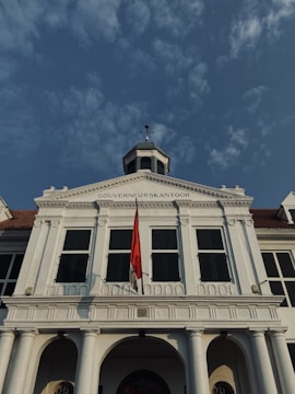 A historic white building with a tall structure, featuring an archway and large windows with green shutters. The building's facade is adorned with columns and a red and white flag is prominently displayed at the front. The sky above is clear with scattered clouds.