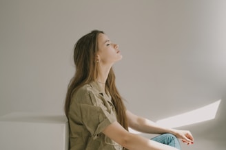 A person with long hair is sitting in profile, eyes closed, in a relaxed and serene posture. The sunlight casts soft shadows against the wall, creating a tranquil atmosphere.