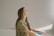 A person with long hair is sitting in profile, eyes closed, in a relaxed and serene posture. The sunlight casts soft shadows against the wall, creating a tranquil atmosphere.