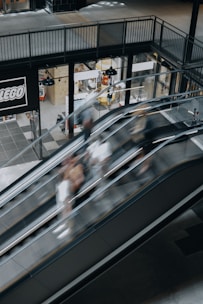 A shopping center setting with escalators showing motion blur of people moving up and down. The surrounding area includes a shop with a prominent LEGO sign, displaying various products through large windows. Industrial-style lighting hangs from the ceiling, and the flooring is a mix of tiles in neutral colors.